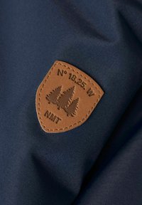 Tumesinine kangas, millel on pruun nahast barett, millel on kilbi kuju, reljeefne puudega ning tekst "N° 18.25 W NMT" allosas.
