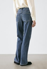 Niebieskie dżinsy z prostą nogawką, pięcioma kieszeniami i widocznym przeszyciem. Noszone z jasnobarwną, długorękawową koszulą.