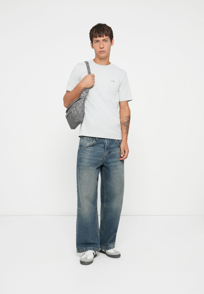 Camiseta gris, jeans azul de corte holgado, bolso bandolera gris. Zapatillas deportivas blancas y grises, diseño minimalista, atuendo casual en tonos neutros.