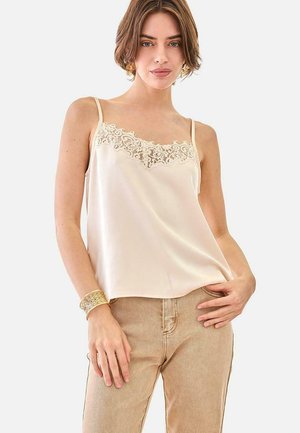 Mujer con camiseta de satén beige con detalles de encaje y pantalones beige, complementada con una pulsera ancha dorada, posando frente a un fondo liso.