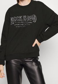 Felpa nera con grafica "ROCK IS DEAD" in argento. Presenta un collo rotondo e una vestibilità ampia, abbinata a pantaloni di pelle nera lucida.