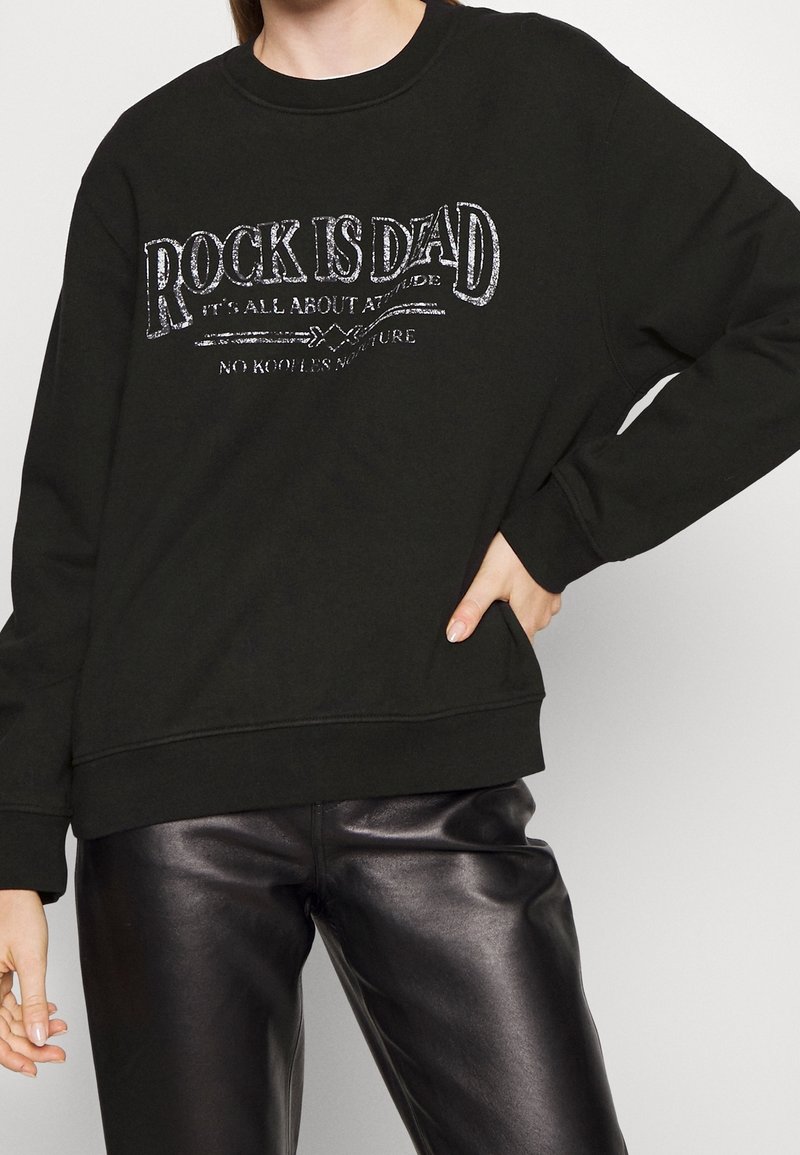 Felpa nera con grafica "ROCK IS DEAD" in argento. Presenta un collo rotondo e una vestibilità ampia, abbinata a pantaloni di pelle nera lucida.