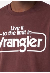 Primo piano di una persona che indossa una t-shirt bordeaux con testo bianco: "Vivi al limite con Wrangler."