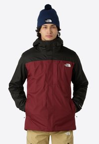 Giacca da uomo con corpo color bordeaux e maniche nere, tessuto impermeabile, cappuccio completo e tasca frontale con zip. Indossata con pantaloni beige e un berretto blu.