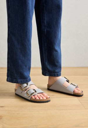 Grå slidesandaler med to justerbare stropper og store sølvspænder, der har en korkfodbund og sål i sort gummi.