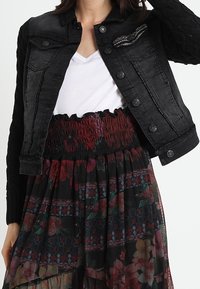 Nainen, joka pitää päällään mustaa farkkutakkia valkoisen V-pääntie-paidan ja korkea vyötäröinen, kukkakuosisen hameen, jossa on rypytetty vyötärö.