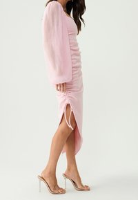 Woman in a light pink ruched midi dress with long puff sleeves and clear high-heeled mules standing on a plain background.