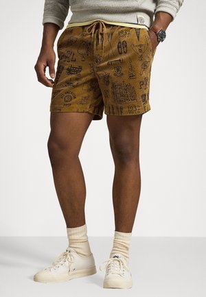 Homme portant un short jaune moutarde avec des croquis noirs, des chaussettes beiges, des baskets blanches, un sweat gris et une montre-bracelet, debout avec une main dans la poche.
