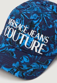 Kapa s plavim cvjetnim uzorkom s bijelim logotipom "VERSACE JEANS COUTURE" u masivnom fontu. Izrađena od teksturiranog materijala, s zakrivljenim vizirima.
