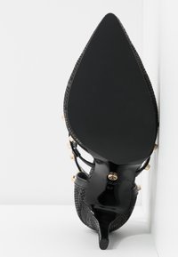 Chaussure à talon noir avec un bout pointu, un dessus texturé et des accents dorés. Elle possède un talon fin et une semelle noire lisse.