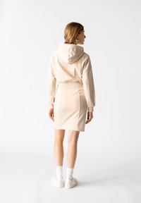 Beige Kapuzensweatshirt mit verkürztem Schnitt, kombiniert mit einem taillierten beigen Rock. Das Model trägt weiße Sneakers mit Socken, die ein einfaches Design zeigen.