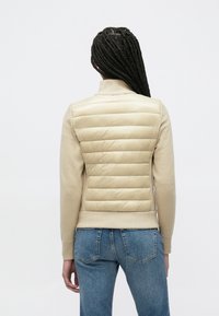 Μπεζ υβριδικό μπουφάν με επενδυμένη, γεμιστή πλάτη, λεία μπροστινή όψη, ψηλό γιακά και ριμπ μανσέτες. Συνδυάζεται με μπλε τζιν παντελόνι.