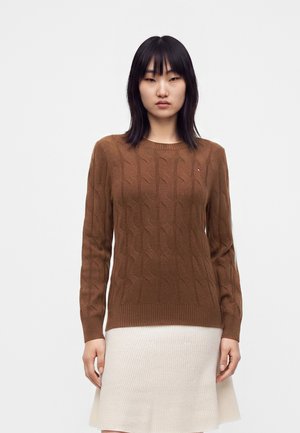 Femme aux cheveux noirs raides portant un pull marron en maille torsadée et une jupe côtelée crème, debout devant un fond blanc uni.