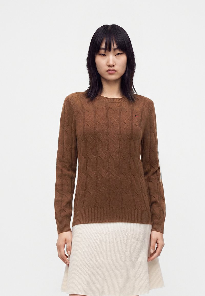 Femme aux cheveux noirs raides portant un pull marron en maille torsadée et une jupe côtelée crème, debout devant un fond blanc uni.