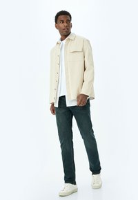 Camisa de botones beige clara con un bolsillo en el pecho, llevada sobre una camiseta blanca, combinada con jeans verde oscuro y zapatillas blancas.