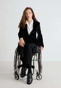 Blazer de terciopelo negro sobre una camisa blanca, combinado con pantalones anchos negros. Persona en una silla de ruedas plateada que lleva zapatos negros brillantes.