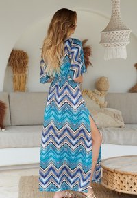 Maxikleid im blauen und braunen Zickzackmuster, mit einem Schlitz an der Seite und lockeren Ärmeln, aus leichtem Stoff gefertigt.