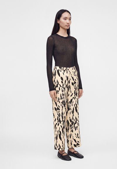Femme debout portant un haut noir transparent à manches longues, un pantalon crème taille haute avec des taches noires irrégulières et des chaussures plates noires.