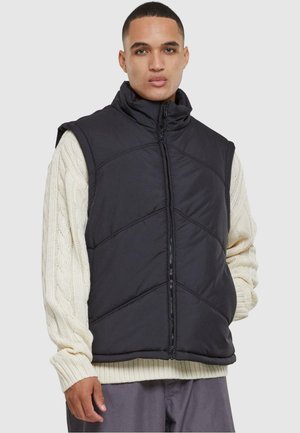 Zwarte geïsoleerde vest met een hoge kraag en ritssluiting. Heeft een gewatteerd ontwerp en een mouwloze snit, gedragen over een crèmekleurige kabelgebreide trui.