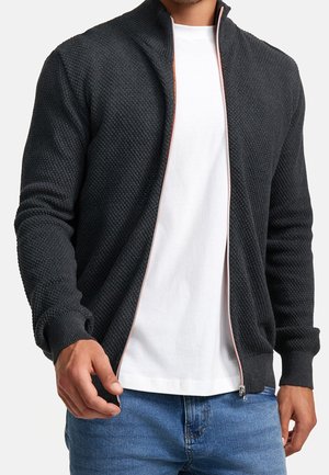 Mężczyzna ubrany w czarny, teksturowany sweter na suwak, na białej koszulce i niebieskich dżinsach, stojący na jednolitym tle.