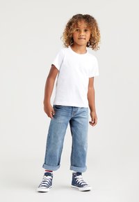 Vit bomullströja, blå denimjeans uppvikta vid fållen och blå högskor med vita sulor. Barnet poserar självsäkert i en avslappnad outfit.