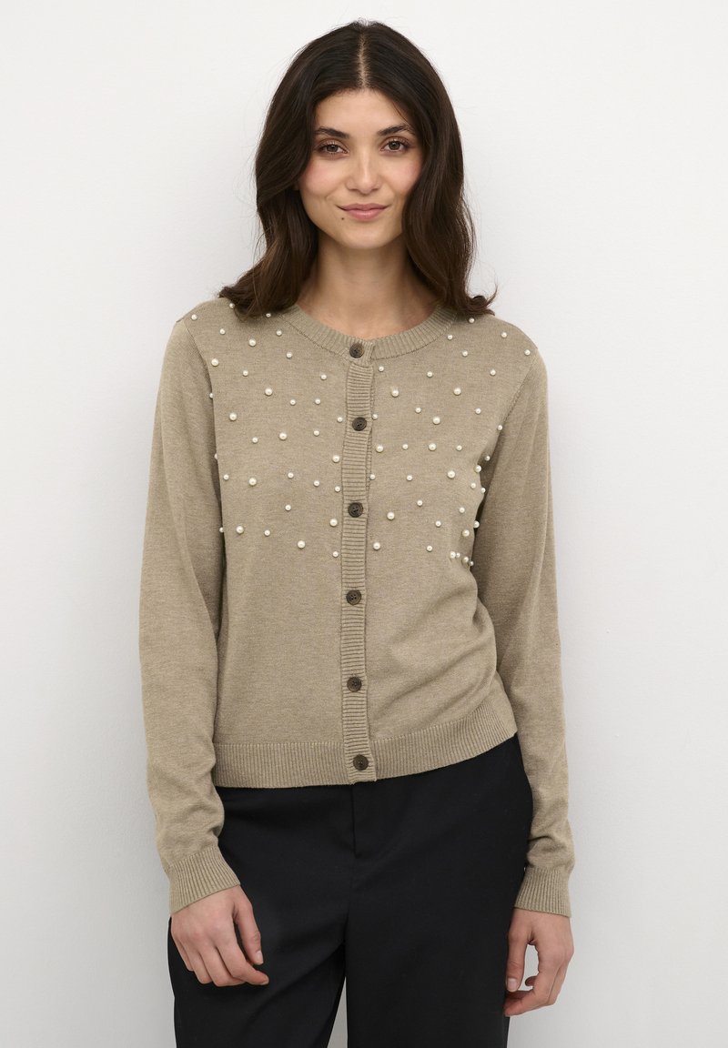Bēšs kardigans ar teksturētu virsmu, kurā ir krēmkrāsas pērles un tumši pogas. Garas piedurknes un ribotas aproces un apakšmalas.