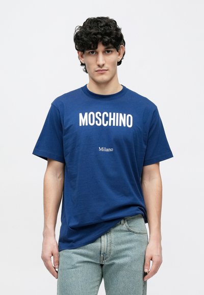 Tamnoplava pamučna majica kratkih rukava s velikim bijelim natpisom "MOSCHINO" i manjim natpisom "Milano" ispod, uparena s laganim trapericama.
