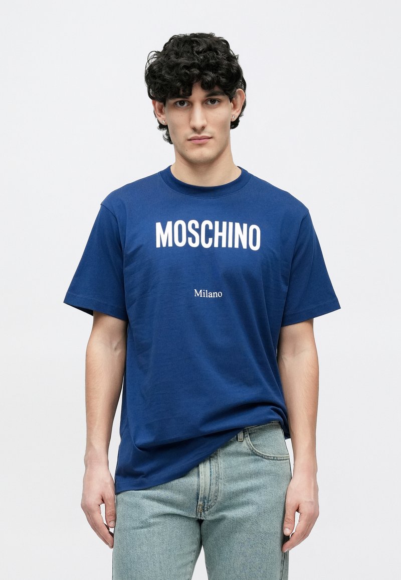 Marineblauw katoenen T-shirt met korte mouwen, met grote witte tekst "MOSCHINO" en daaronder kleinere tekst "Milano", gecombineerd met lichte spijkerbroek.