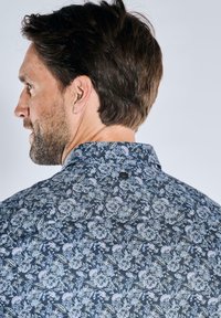 Dunkelblaues Hemd mit floralen Mustern und hellen Blumen. Hat einen spitzen Kragen und ein kleines Logo am hinteren Halsauschnitt.