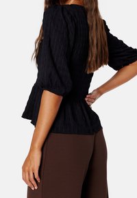 Blouse noire texturée avec manches bouffantes et un design évasé style péplum, associée à un pantalon marron ajusté. Vue de côté mettant en valeur la texture du tissu.