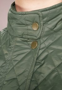 Grüne gesteppte Jacke mit hohem Kragen, zwei Messingknöpfen und diagonaler Steppnaht. Strukturierter, glatter Stoff.