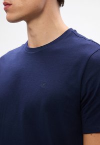 Homme portant un t-shirt bleu marine uni à col rond avec un petit logo en relief "cK" sur la poitrine, cadré de l'épaule au menton.