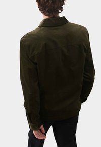 Ciemnozielona kurtka z korduroy z kołnierzem, zapięciem na guziki i prostym krojem. Tekstura jest żeberkowana; minimalny widoczny szew.