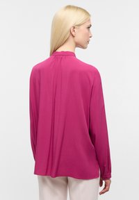 Blusa a maniche lunghe di un vivace fucsia, realizzata in tessuto morbido, con polsini con bottoni e un collo alto con sottili pieghe sulla parte posteriore.