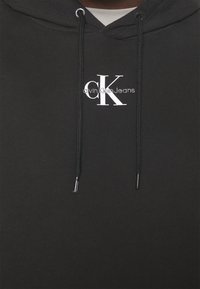 Čierna mikina s kapucňou s šnúrkou so striebornými agletmi, vyrazené biele logo Calvin Klein Jeans na hrudi, bavlnený materiál, ležérny dizajn.