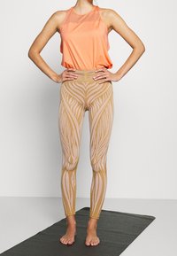 Träningsklädsel bestående av en ventilerande orange linne och mönstrade beige leggings med djärva, spiralformade mönster i ljusa och mörka nyanser.