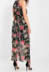 Kvinna iklädd en ärmlös, ankellång svart klänning med röd och vit blommig tryck, tillsammans med svarta högklackade sandaler, stående med ryggen vänd.