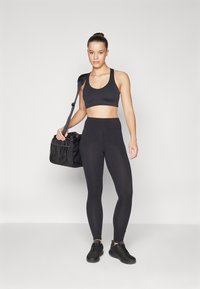 Top deportivo negro y leggings de talle alto, ambos en un material elástico. Se combinan con zapatillas negras y se sostiene una mochila de gimnasio negra. Diseño minimalista.