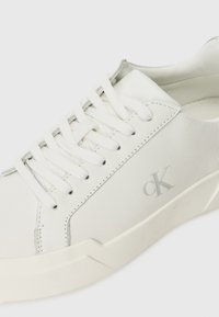 Weiße Leder-Sneaker mit einer strukturierten Oberfläche, minimalistischen Design, flachen Schnürsenkeln und einem dezenten grauen Logo an der Seite.