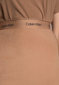 Hnědý top Calvin Klein s odpovídajícím elastickým pasem, na kterém je v černém textu uvedeno jméno značky, vyrobený z měkké, texturované tkaniny.