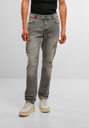 Szürke farmernadrág, slim fit fazonnal, kopott részletekkel és fehér festékcseppekkel. A hátsó zseb előtt piros részlet található.