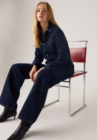 Denim jumpsuit i mørkeblå med lange ærmer, knaplukning foran og brystlomme. Modellen sidder på en kromstol med en rød sæde.