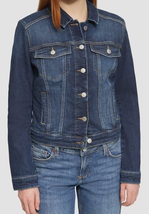 Femme portant une veste en jean bleu avec des boutons argentés par-dessus un jean bleu, montrant le haut du corps des épaules aux hanches sur un fond uni.