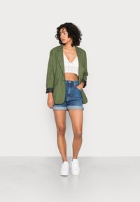 Blazer xadrez verde com detalhes pretos, top cropped de renda branca, calções de ganga azuis de cintura alta e ténis brancos. Conjunto simples e casual.