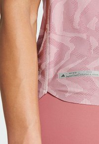 Top deportivo de color rosa claro con un patrón texturizado, que presenta rayas blancas y una etiqueta del logo de Adidas en el dobladillo, mostrando un diseño ajustado.