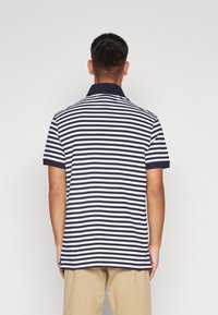 Muž oblečený v námořnicky modrém a bílém horizontálně pruhovaném polo tričku s krátkým rukávem a béžových kalhotách, stojící čelem k jednoduchému bílému pozadí.