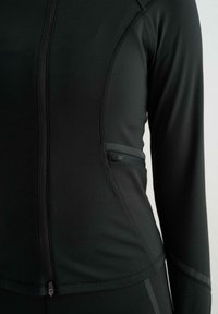 Černá sportovní bunda se zipem, vyrobená z hladkého, pružného materiálu a s plochými švy. Obsahuje boční kapsu s vroubkovaným akcentem.