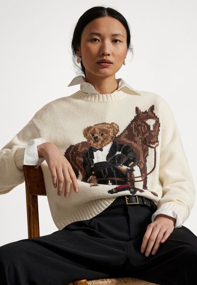 Kobieta siedząca, ubrana w kremowy sweter z wzorem przedstawiającym misia w smokingu, obok brązowego konia, czarne spodnie i biała koszula z kołnierzykiem.