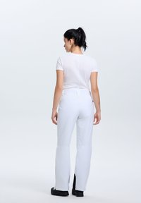 Pantaloni da sci bianchi con una vestibilità su misura, dotati di aperture laterali con zip e una texture liscia. Indossati con un top bianco aderente e calzature nere.