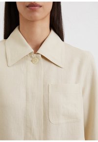 Beige linnen shirt met een grote kraag, een enkele knoopsluiting en een patchzak aan de linkerkant. Gestructureerde stof met een gladde afwerking.